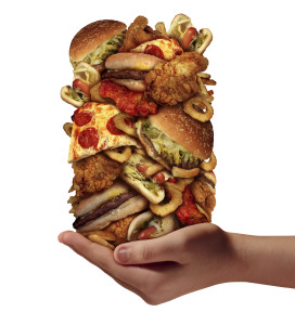Over eating and compulsive indulgence of fast food concept as a hand holding up a huge stack of junk food as hamburgers hotdogs and french fries as an unhealthy diet nd bad nutrition symbol isolated on a white background. Photo: wildpixel/iStock